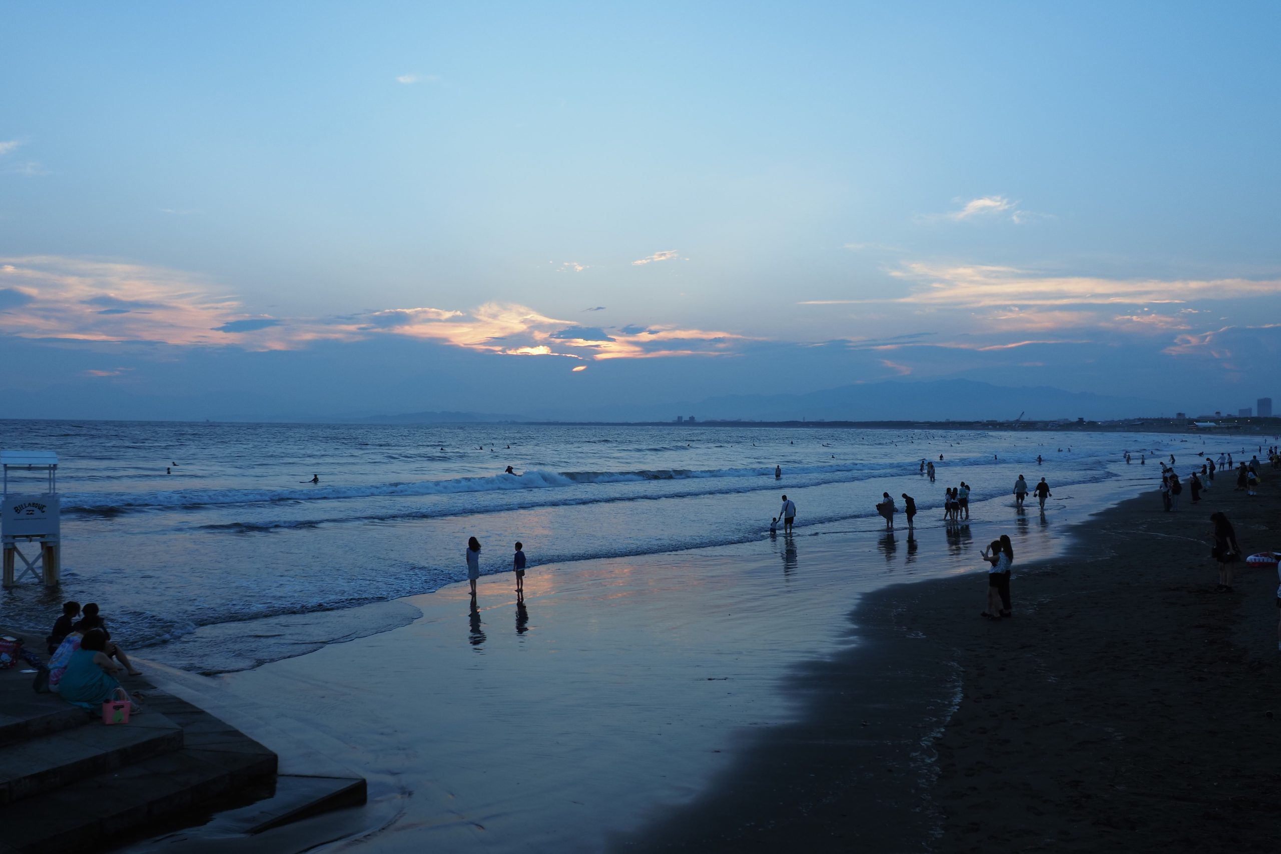 淡い青と夕焼けに染まる海辺と波打ち際の人々